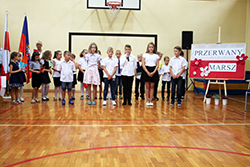 Grupa dzieci w trakcie uroczystej akademmi przedstawia program artystyczny. Dzieci mówia do mikrofonów. Są na galowo ubrane.