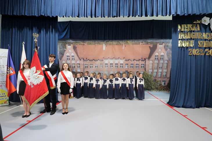 Odświętnie ubrane dzieci stoją na baczność, by odśpiewać hymn. Jest również poczet sztandarowy