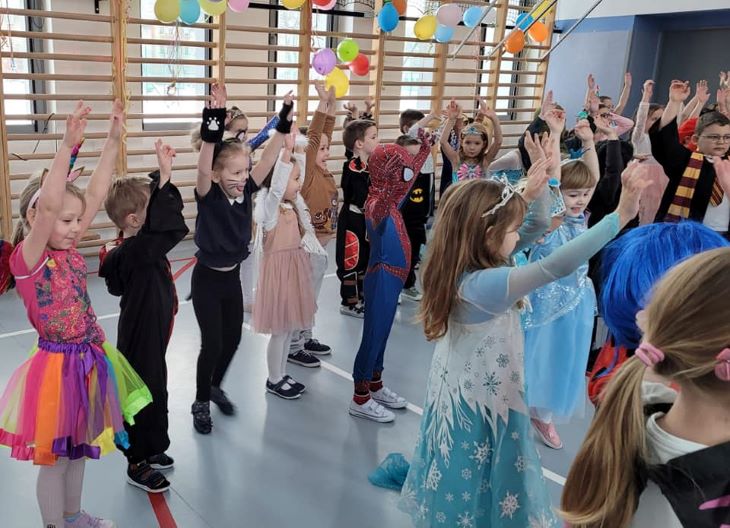 Dzieci przebrane w różne stroje karnawałowe bawią się w sali udekorowanej balonami.