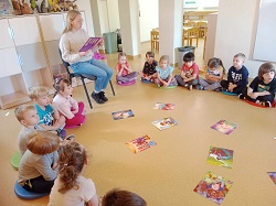 dzieci siedzą w kole na podłodze, w środku koła leżą książki, dorosła osoba czyta dzieciom bajkę