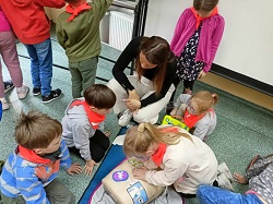 na zdjęciu dzieci w kole stoją lub siedzą dookoła fantomu do pierwszej pomocy