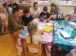 na zdjęciu dzieci z swoimi rodzicami siedzą dookoła stolików na których przeprowadzają doświadczenia 