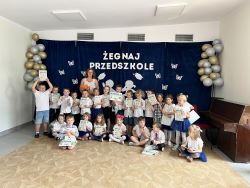 Grupa dzieci stoi z panią na tle dekoracji "Żegnaj przedszkole". Dzieci ubrane są w galowe stroje i trzymają w rękach książki 