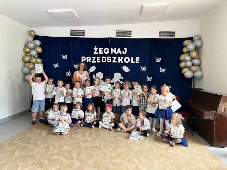 Grupa dzieci stoi z panią na tle dekoracji "Żegnaj przedszkole". Dzieci ubrane są w galowe stroje i trzymają w rękach książki 