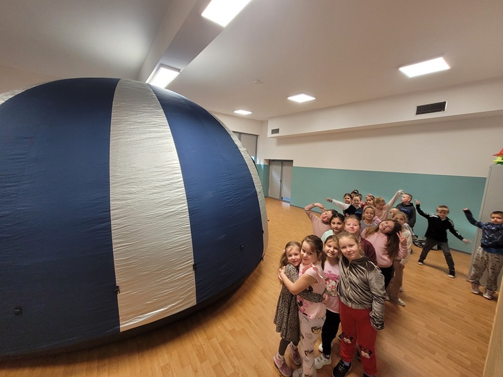 Na zdjęciu widać grupę dzieci stojącą obok dużego, nadmuchiwanego obiektu w kształcie kopuły. Jest to przenośne planetarium ustawione w sali gimnastycznej lub szkolnej. Dzieci wyglądają na podekscytowane i gotowe do uczestnictwa w pokazie. Wnętrze sali jest przestronne, z jasnymi ścianami i podłogą z drewnianych paneli.