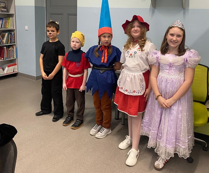 Na zdjęciu widać grupę pięciorga dzieci stojących w szeregu, ubranych w kolorowe stroje bajkowych postaci 🎭. Znajdują się w sali szkolnej, prawdopodobnie w bibliotece – w tle widać regał z książkami