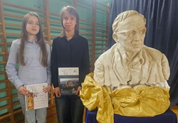 Na zdjęciu widać dwie dziewczyny stojące obok siebie w sali gimnastycznej, przy dużym popiersiu przedstawiającym starszego mężczyznę w okularach. Dziewczyny trzymają w rękach książki oraz dyplomy, co sugeruje, że mogły otrzymać nagrody lub wyróżnienia w konkursie szkolnym.