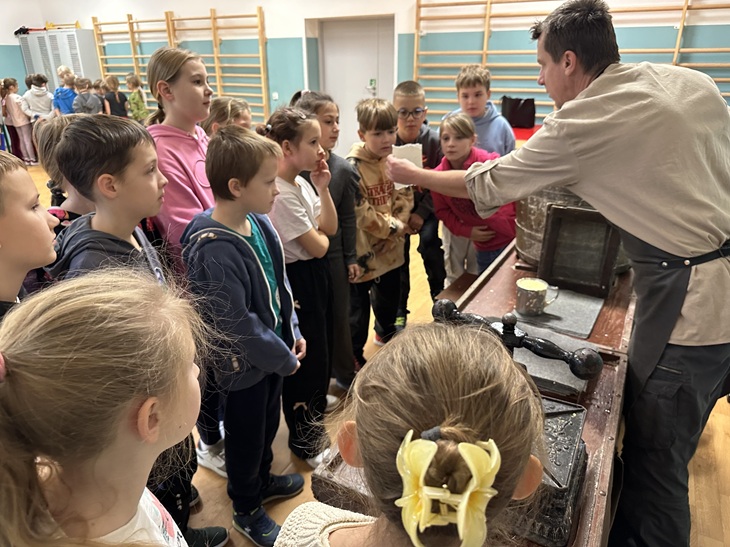 Dzieci stoją przy stole rzemieślniczym, a prowadzący pokazuje im papier ręcznie czerpany. Są to warsztaty tworzenia papieru.