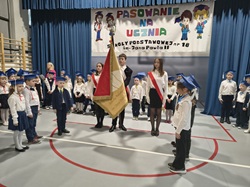 Na zdjęciu widać uroczystość pasowania na ucznia w szkole podstawowej. W centrum stoi poczet sztandarowy: trzech uczniów trzymających szkolny sztandar, ubranych elegancko — chłopiec w garniturze trzyma sztandar, a dwie dziewczyny w białych koszulach i czarnych spódnicach stoją po jego bokach z biało-czerwonymi szarfami.