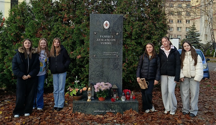 Na zdjęciu widoczna jest grupa sześciu młodych osób stojących po obu stronach pomnika Pamięci Zesłańców Sybiru. Pomnik jest wykonany z ciemnego kamienia, ustawiony na tle gęstych, zielonych krzewów. U jego podstawy znajdują się zapalone znicze, kwiaty oraz drobne pamiątkowe dekoracje, co tworzy atmosferę zadumy i szacunku.