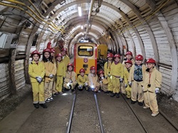 Na zdjęciu widać grupę dzieci ubranych w żółte kombinezony ochronne i czerwone kaski górnicze. Stoją one w tunelu kopalnianym, obok małej górniczej kolejki o żółto-czerwonych barwach. W tle widać oświetlony korytarz z metalowymi obudowami i żółtymi przewodami biegnącymi wzdłuż ścian.