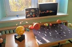 Na zdjęciu widać szkolną wystawę przestrzennych modeli Układu Słonecznego, wykonanych przez uczniów w ramach konkursu. Prace ustawione są na ławkach i parapecie w klasie, oświetlonej naturalnym światłem z okna. Na pierwszym planie znajduje się model z dużym żółtym Słońcem i orbitującymi wokół niego planetami umocowanymi na drucikach lub patyczkach. Obok widoczny jest większy projekt z czarną podstawą ozdobioną gwiazdami, na której rozmieszczono kolorowe kule symbolizujące planety – każda z nich jest podpisana.