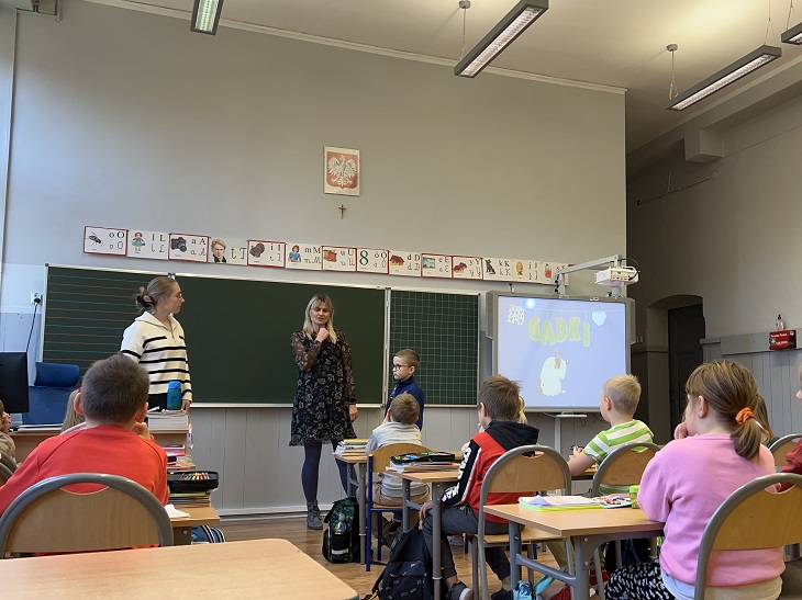 Na zdjęciu widać zajęcia prowadzone w klasie szkolnej. Przy tablicy stoją dwie osoby dorosłe – prawdopodobnie pedagog i psycholog – które rozmawiają z uczniami. Jedno z dzieci uczestniczy aktywnie w zajęciach, stojąc przy prowadzących, podczas gdy pozostali uczniowie siedzą w ławkach i uważnie słuchają.