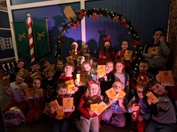 Na zdjęciu widać grupę dzieci pozujących razem w świątecznej scenerii, z dekoracjami bożonarodzeniowymi i świetlną girlandą. Dzieci trzymają dyplomy lub kartki i wyglądają na zadowolone.