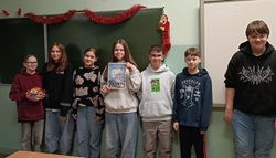 Na zdjęciu widać grupę uczniów stojących w klasie szkolnej. Pozują razem przed tablicą, jeden z uczniów trzyma dyplom, a całość sugeruje podsumowanie lub wręczenie nagród po konkursie. Atmosfera jest radosna i swobodna.