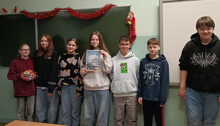 Na zdjęciu widać grupę uczniów stojących w klasie szkolnej. Pozują razem przed tablicą, jeden z uczniów trzyma dyplom, a całość sugeruje podsumowanie lub wręczenie nagród po konkursie. Atmosfera jest radosna i swobodna.