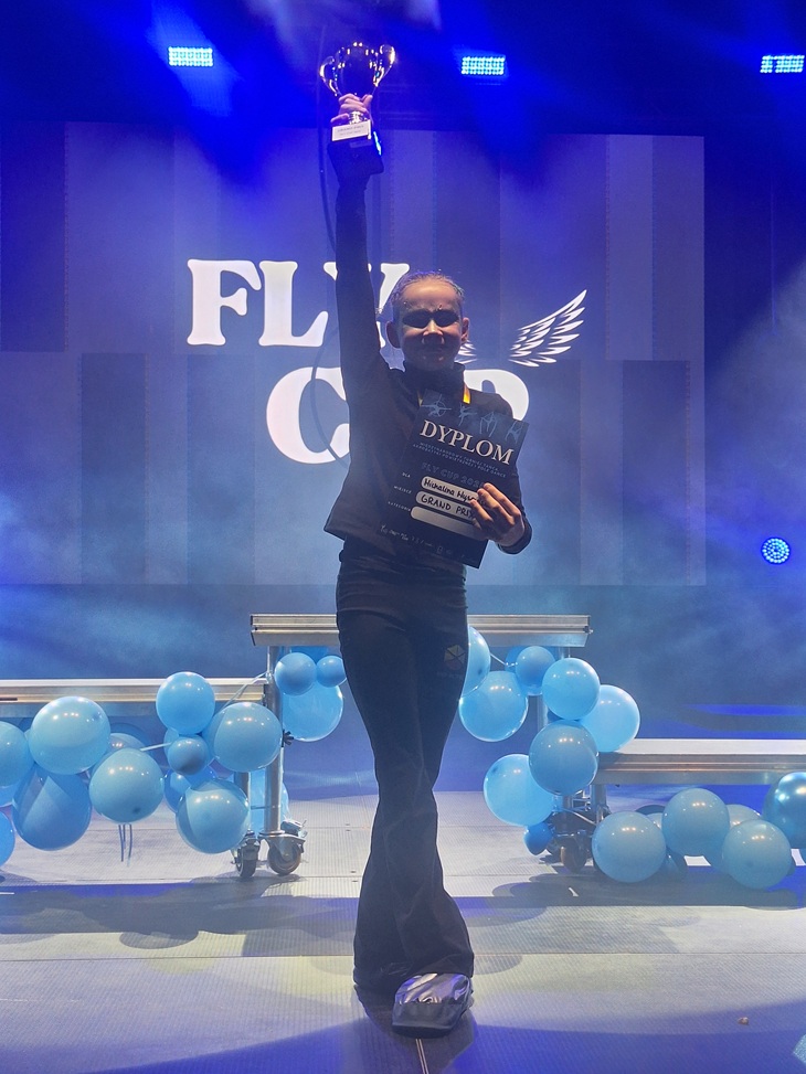 Na zdjęciu widać młodą dziewczynę stojącą na scenie podczas uroczystej gali lub zawodów. Trzyma wysoko uniesiony puchar oraz dyplom, co świadczy o jej zwycięstwie. Za nią znajduje się duże, podświetlone logo „FLY CUP”. Scena jest oświetlona kolorowymi światłami, a po obu stronach umieszczone są dekoracje z niebieskich balonów. Dziewczyna wygląda na dumną i szczęśliwą z osiągniętego sukcesu.