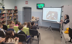 Na zdjęciu widać grupę dzieci siedzących w szkolnej bibliotece i uczestniczących w zajęciach. Przed nimi stoi nauczycielka prowadząca prezentację o pluszowych misiach, wyświetlaną na ekranie projektora. W tle znajdują się regały z książkami oraz pluszowe misie ustawione na stoliku.