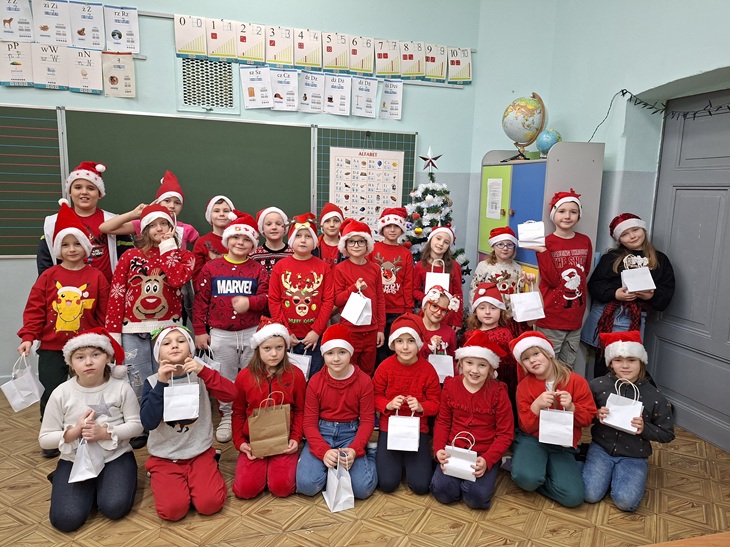 Na zdjęciu widać dużą grupę dzieci pozujących razem w klasie podczas mikołajkowego świętowania. Wszystkie dzieci mają na głowach czerwone czapki Mikołaja, a większość z nich jest ubrana w czerwone lub świąteczne swetry z motywami reniferów, Mikołaja czy zimowych wzorów. Trzymają w rękach małe, jednakowe paczuszki – wyglądają jak upominki rozdane z okazji Mikołajek.  W tle znajduje się tablica szkolna, dekoracje oraz mała choinka ustawiona na szafce. Atmosfera na zdjęciu jest radosna i pełna świątecznego klimatu, a dzieci uśmiechają się i wyglądają na bardzo zadowolone.