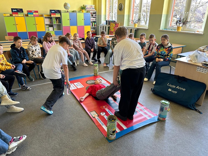 Na zdjęciu widać klasę szkolną, w której odbywają się zajęcia z pierwszej pomocy. Uczniowie siedzą w kręgu, uważnie obserwując praktyczny pokaz.  Na środku sali leży mata edukacyjna z ilustracjami i instrukcjami dotyczącymi udzielania pierwszej pomocy. Dwóch uczniów demonstruje czynności ratownicze na osobie leżącej na macie — wygląda to na ćwiczenia związane z ewakuacją lub ułożeniem poszkodowanego.