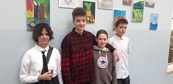 Na zdjęciu widać czworo młodych uczniów stojących obok siebie na szkolnym korytarzu. W tle wiszą kolorowe prace plastyczne. Osoba po lewej ma krótkie, kręcone włosy, jest ubrana w białą koszulę i czarny krawat. Obok stoi wysoka osoba w czerwono-czarnej koszuli w kratę, z lekkim uśmiechem. Trzecia osoba ma na sobie jasną bluzę z kapturem i stoi bardzo blisko poprzedniej osoby, obejmując ją ramieniem. Po prawej stronie stoi czwarta osoba w białej koszuli, patrząca lekko w bok.