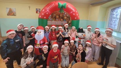 Na zdjęciu widać grupę dzieci w świątecznych czapkach Mikołaja, pozujących w sali szkolnej. W tle znajdują się bożonarodzeniowe dekoracje, dmuchana brama z napisem „Merry Christmas” oraz postacie Mikołaja i bałwana. Atmosfera jest radosna i wyraźnie świąteczna.