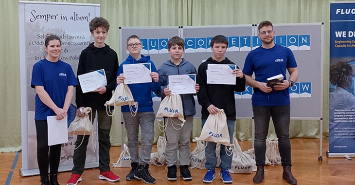 Na zdjęciu widzimy grupę młodych ludzi oraz dwóch dorosłych, którzy biorą udział w wydarzeniu związanym z konkursem promującym rozwój umiejętności STEM (nauka, technologia, inżynieria, matematyka). Uczestnicy konkursu trzymają certyfikaty i torby z nagrodami.  W tle widnieją banery z logo konkursu „Fluo Competition”
