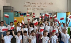 Na zdjęciu widać grupę dzieci w sali lekcyjnej, ubrane w białe koszulki z przypiętymi kokardkami narodowymi. Każde z dzieci trzyma w ręce małą polską flagę. W tle, na ścianie, widnieje cytat Jana Pawła II:  „Wymagajcie od siebie, choćby inni od was nie wymagali.” — Jan Paweł II