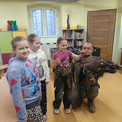 Na zdjęciu widzimy trzy uśmiechnięte dziewczynki stojące obok mężczyzny, który trzyma na rękawicy dużego ptaka drapieżnego – orła 🦅. Dzieci z zainteresowaniem i radością obserwują ptaka, a jedna z dziewczynek delikatnie go dotyka. Zdjęcie zostało wykonane w szkolnej sali, co widać po otoczeniu – są tam regały z książkami, stół do gry w piłkarzyki oraz kolorowe szafki. To najprawdopodobniej moment z opisanego wcześniej spotkania edukacyjnego z orłem.