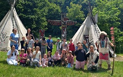 Na zdjęciu widać grupę dzieci i opiekunów pozujących do wspólnej fotografii w Wiosce Indiańskiej. W tle znajdują się charakterystyczne tipi oraz kolorowy totem, a obok grupy stoi osoba przebrana za rdzennych mieszkańców Ameryki Północnej, ubrana w tradycyjny strój z pióropuszem i laską ozdobioną piórami.
