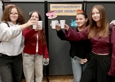 Na zdjęciu widać cztery uczennice stojące obok siebie, które wznoszą toast papierowymi kubeczkami z herbatą. Dziewczęta są uśmiechnięte, a cała scena oddaje miłą i radosną atmosferę wspólnego świętowania.  W tle znajduje się gazetka tematyczna z napisem „Międzynarodowy Dzień Herbaty” oraz dekoracje nawiązujące do tego wydarzenia, co wskazuje, że zdjęcie zostało wykonane podczas szkolnych obchodów tego święta. Całość podkreśla integrację uczniów oraz przyjemny charakter spotkania.