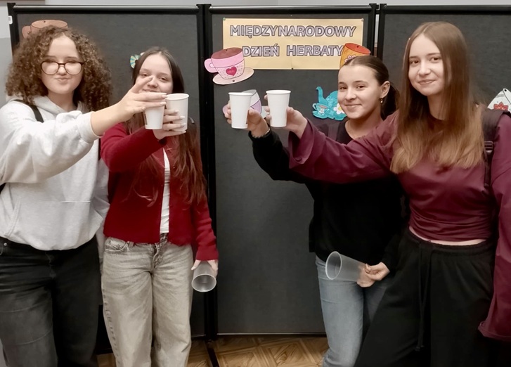 Na zdjęciu widać cztery uczennice stojące obok siebie, które wznoszą toast papierowymi kubeczkami z herbatą. Dziewczęta są uśmiechnięte, a cała scena oddaje miłą i radosną atmosferę wspólnego świętowania.  W tle znajduje się gazetka tematyczna z napisem „Międzynarodowy Dzień Herbaty” oraz dekoracje nawiązujące do tego wydarzenia, co wskazuje, że zdjęcie zostało wykonane podczas szkolnych obchodów tego święta. Całość podkreśla integrację uczniów oraz przyjemny charakter spotkania.