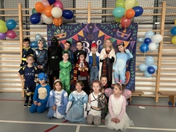 Na zdjęciu widać grupę uśmiechniętych dzieci w kolorowych strojach karnawałowych. Przebrane za bohaterów bajek, księżniczki, rycerzy oraz postacie z filmów, pozują na sali gimnastycznej na tle dekoracji balowej i balonów.