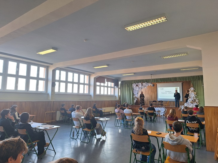 Na zdjęciu widać uczniów siedzących przy stolikach w sali szkolnej, uczestniczących w konkursie. Z przodu sali znajduje się scena z prowadzącymi oraz prezentacją multimedialną. W sali panuje atmosfera skupienia i pracy.