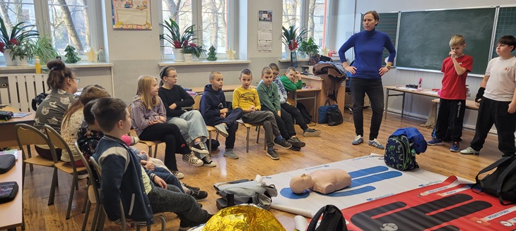Zdjęcie przedstawia scenę z sali lekcyjnej, w której odbywa się praktyczne szkolenie z zakresu pierwszej pomocy. Grupa dzieci siedzi w półokręgu, uważnie słuchając dorosłego instruktora stojącego przy tablicy. Na podłodze znajdują się maty treningowe oraz fantom do nauki resuscytacji krążeniowo-oddechowej (CPR), a także inne akcesoria ratownicze, takie jak koc termiczny i torby medyczne.