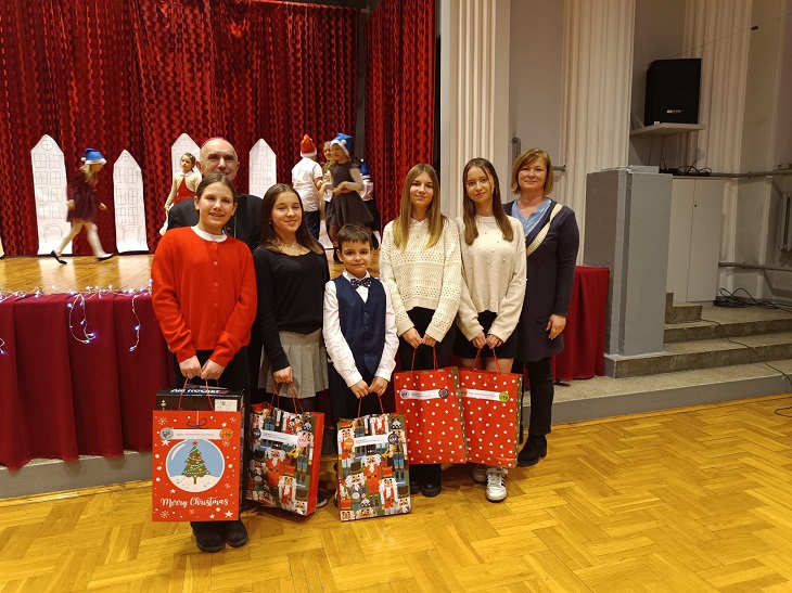 Uczniowie z nauczycielka stoja na tle sceny, w rękach trzymaja torby z upominkami, są odświętnie ubrani