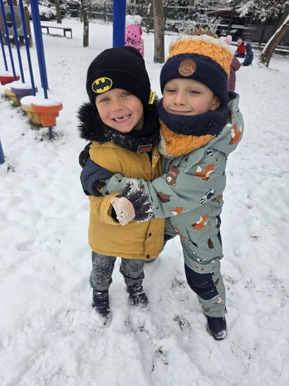 dwóch chłopców  w kombinezonach z uśmiętymi buziami na podwórku pokrytym śniegiem