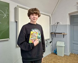 Na zdjęciu widać ucznia stojącego w sali lekcyjnej, który trzyma w ręku pudełko gry planszowej „Chińczyk”. Fotografia przedstawia zwycięzcę szkolnego turnieju podczas wręczenia nagrody.