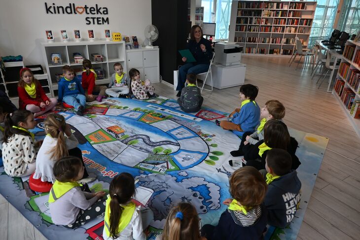 dzieci siedzące na dywanie i słuchające z zaciekawieniem co mówi prowadząca zajęcia edukacyjne