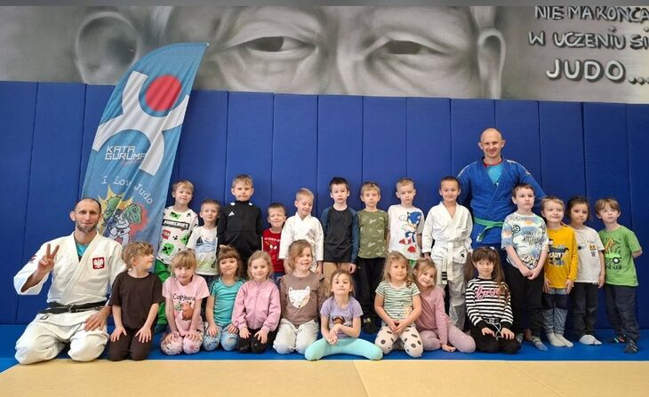 grupa dzieci ubrana w sportrowe stroje stojąca na sali gimnastycznej z trenerami judo ubranymi w szaty do judo.