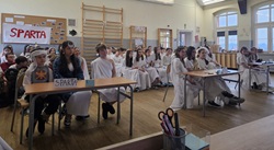 Na zdjęciu widać uczniów siedzących w sali lekcyjnej podczas konkursu tematycznego. Większość dzieci jest przebrana w stroje stylizowane na starożytną Grecję — białe tuniki i opaski na głowie. Na pierwszym planie znajduje się stolik z napisem „Sparta”, a w tle widać plakat z tą samą nazwą. Uczniowie siedzą w drużynach, uważnie słuchając lub przygotowując się do odpowiedzi, co podkreśla konkursową atmosferę wydarzenia.