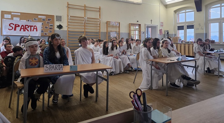 Na zdjęciu widać uczniów siedzących w sali lekcyjnej podczas konkursu tematycznego. Większość dzieci jest przebrana w stroje stylizowane na starożytną Grecję — białe tuniki i opaski na głowie. Na pierwszym planie znajduje się stolik z napisem „Sparta”, a w tle widać plakat z tą samą nazwą. Uczniowie siedzą w drużynach, uważnie słuchając lub przygotowując się do odpowiedzi, co podkreśla konkursową atmosferę wydarzenia.