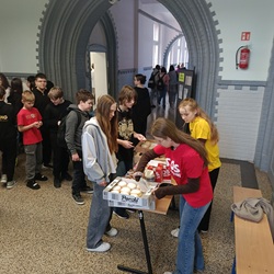 Plakat promuje szkolną akcję charytatywną z okazji Tłustego Czwartku, która odbędzie się 12 lutego pod hasłem „Wyślij pączka do Afryki”. W centralnej części znajduje się zdjęcie pączków z logo akcji. Całość utrzymana jest w jasnej kolorystyce, ozdobiona czerwonymi i różowymi sercami oraz ilustracjami pączków. Na dole plakatu podano harmonogram sprzedaży: o 9:40 i 10:35 na parterze oraz I i II piętrze, a także o 11:30 i 12:30 na I piętrze.