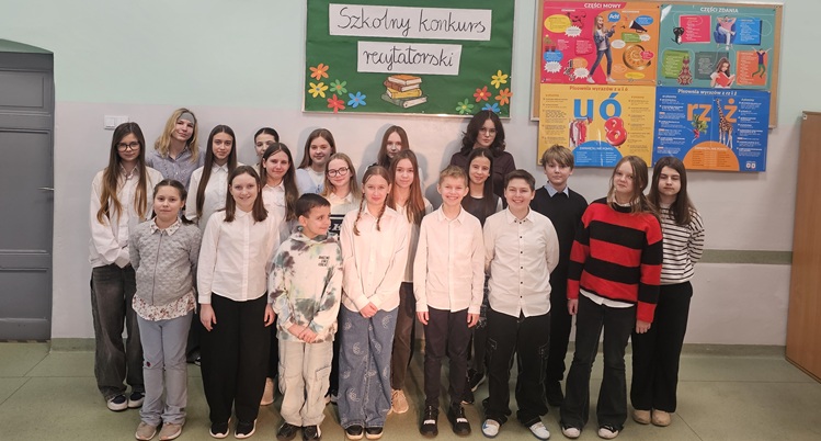 Na zdjęciu widać grupę młodzieży szkolnej stojącej w szkolnym korytarzu. Uczniowie ustawieni są w kilku rzędach, ubrani odświętnie – dominują białe koszule i ciemne spodnie. Za nimi znajduje się tablica z napisem „Szkolny konkurs recytatorski” oraz kolorowe plakaty edukacyjne. Grupa pozuje do wspólnego, pamiątkowego zdjęcia.