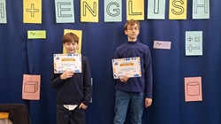 Na zdjęciu widać dwóch uczniów stojących na tle szkolnej gazetki tematycznej związanej z matematyką i językiem angielskim („Math + English”). Chłopcy trzymają dyplomy potwierdzające zdobycie III miejsca w konkursie matematycznym w języku angielskim. W tle znajdują się kolorowe plansze z symbolami matematycznymi i bryłami, podkreślające edukacyjny charakter wydarzenia.