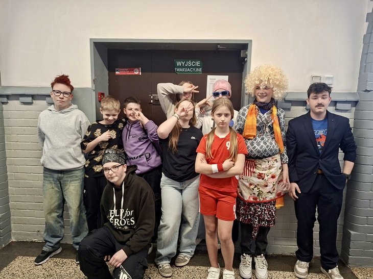 Grupa uczniów stoi na szkolnym korytarzu, przebrana za różne postacie i celebrytów. Kolorowe stroje, peruki i dodatki nadają scenie humorystyczny i kreatywny charakter. Wszyscy pozują do zdjęcia w swobodnej, radosnej atmosferze.
