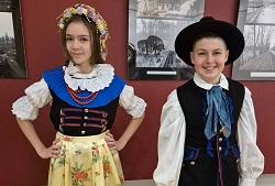 Na zdjęciu widać dwoje uczniów prezentujących tradycyjne stroje śląskie.  Dziewczynka ma na sobie kolorowy strój ludowy z białą bluzką, ozdobnym gorsetem, koralami oraz kwiatowym wiankiem na głowie. Jej strój uzupełnia jasna spódnica z kwiatowym wzorem.  Chłopiec jest ubrany w męski strój śląski – białą koszulę, czarną kamizelkę, kapelusz oraz ozdobną chustę pod szyją.