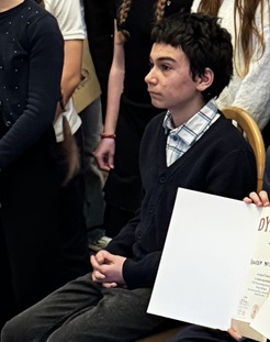 Na zdjęciu widać młodego chłopca siedzącego na krześle podczas uroczystości. Ma na sobie elegancki strój – koszulę w kratę, krawat oraz ciemny sweter. Obok znajduje się dyplom, a w tle stoją inne osoby, prawdopodobnie uczestnicy wydarzenia. Chłopiec wygląda poważnie i skupienie, jak podczas oficjalnego wręczenia nagród.