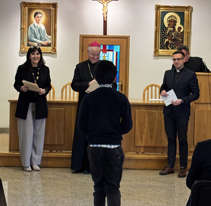 Na zdjęciu widać moment wręczenia nagrody podczas uroczystości. Uczeń podchodzi do stołu, przy którym stoją organizatorzy konkursu, w tym duchowni oraz przedstawicielka szkoły. W tle znajdują się religijne obrazy i krzyż, co wskazuje na uroczysty charakter wydarzenia związanego z konkursem wiedzy biblijnej