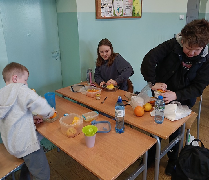 Na zdjęciu uczniowie podczas zajęć w klasie przygotowują zdrowe napoje. Kroją i wyciskają cytrusy – pomarańcze i cytryny – korzystając z wyciskarek i pojemników ustawionych na ławkach. Zajęcia mają praktyczny charakter i pokazują, jak w prosty sposób przygotować świeży, naturalny napój.